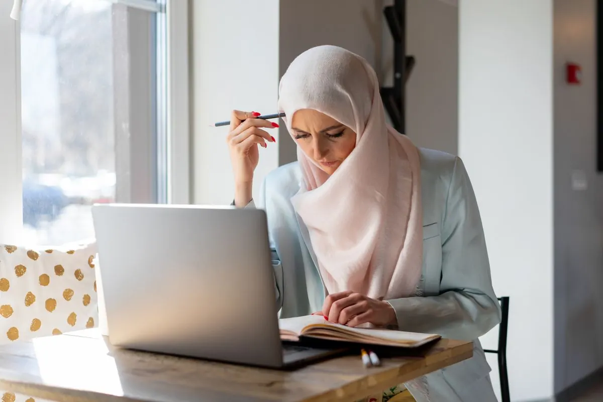 A woman studies an open book beside her laptop.