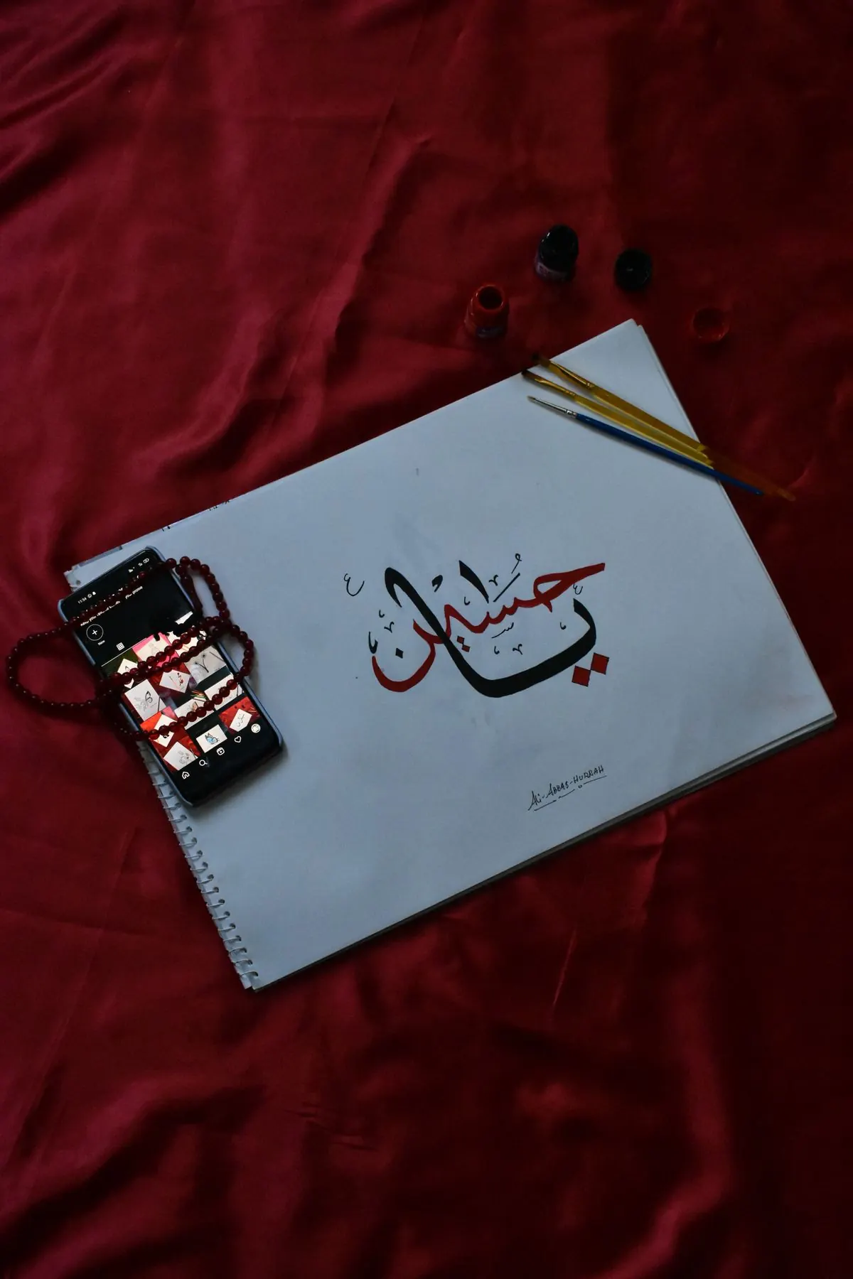 Handwritten Arabic calligraphy in a sketchbook beside brushes and a phone.