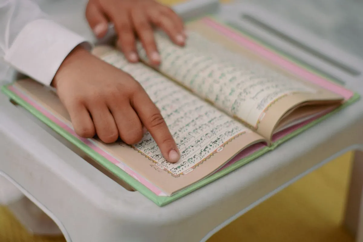 A child follows Quranic text with a finger while the book rests on a desk.