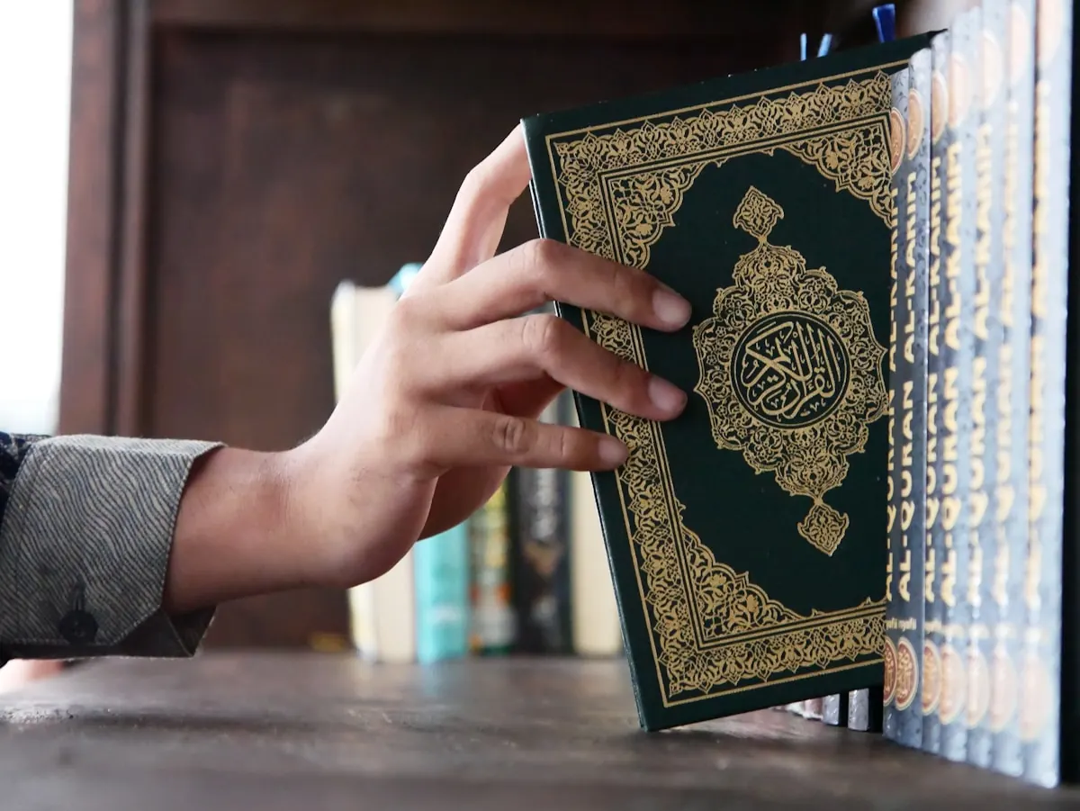 A hand takes a Quran from a wooden bookshelf.