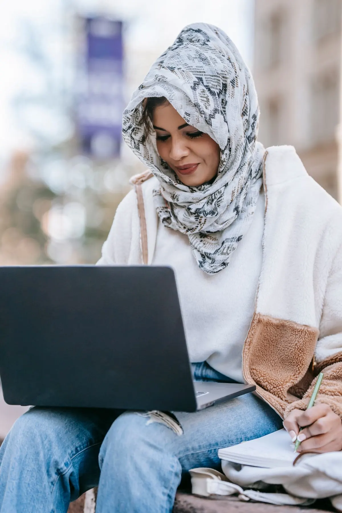 A woman smiles while using a laptop and taking notes outdoors.